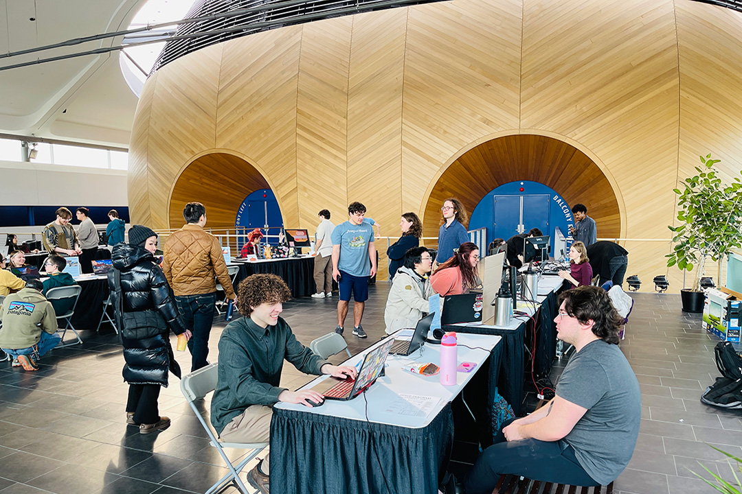 Students sit across from each other at tables in a college atrium where laptops are setup for game playtesting
